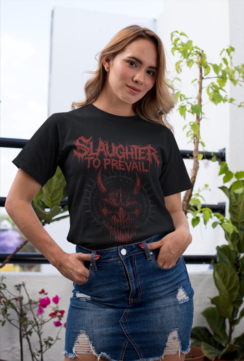 May include: Black t-shirt with the text "SLAUGHTER TO PREVAIL" in red, above a red demonic face graphic. The model is wearing a denim skirt and is standing outside.