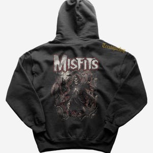 May include: Dark grey hoodie with a graphic of a grim reaper and the word "Misfits" in red and white. The design includes a graveyard scene with a castle, bats, and skulls. The hoodie has a drawstring hood.