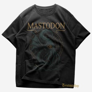May include: Dark gray t-shirt featuring the band name "MASTODON" in gold lettering above a detailed illustration of a sea creature with a large eye and tentacles. The design includes a circular background and water elements.