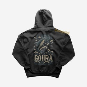 Op de afbeelding: Donkergrijze hoodie met een walvisillustratie, oceaangolven en het woord "GOJIRA". Het ontwerp gebruikt blauwe, witte en grijze tinten. De hoodie heeft een capuchon en een kangoeroezak.
