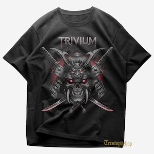May include: Black t-shirt featuring a skull design with a samurai helmet and crossed swords. The word "TRIVIUM" is printed above the skull in a stylized font. The design has red accents and a detailed, graphic style.