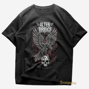 May include: Black t-shirt featuring a graphic design with the text "ALTER BRIDGE" above a raven with outstretched wings perched on a skull. The design includes a decorative floral pattern and the text "ONE DAY REMAINS".