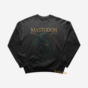 Puede incluir: Sudadera gris oscuro con la palabra "MASTODON" en dorado sobre un gráfico de criatura marina verde azulado y dorado. El diseño incluye un fondo tipo brújula. La sudadera tiene cuello redondo. El nombre de la tienda "Teestopishop" está en la parte inferior derecha.