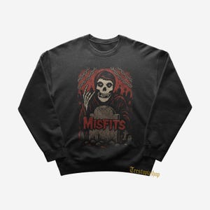 May include: Dark grey sweatshirt with a red and black graphic of a skeleton in a graveyard, the word "MISFITS" is displayed. The design includes chains and tombstones, creating a spooky theme.