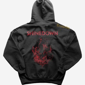 Pode incluir: Moletom cinza escuro com o nome da banda SHINEDOWN em letras vermelhas. Abaixo do texto, há um gráfico vermelho de chamas e fumaça. O moletom tem capuz com cordão e um bolso frontal.