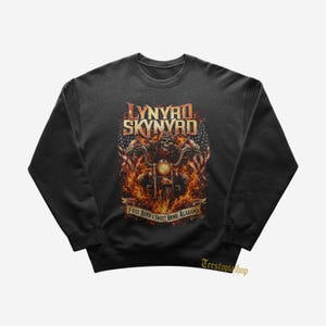 May include: A black crewneck sweatshirt featuring a graphic of a skeleton riding a motorcycle surrounded by flames and American flags. The text reads Lynyrd Skynyrd and Free Bird & Sweet Home Alabama. The Teestopiashop logo is visible in the bottom right.