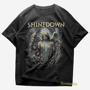 May include: Black t-shirt featuring the band name "SHINEDOWN" in a beige font above a detailed graphic of an angel statue with large wings. The angel has a blue glowing line down its chest. The shirt has a crew neck.