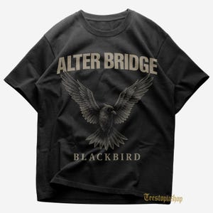 May include: Black t-shirt featuring the text "ALTER BRIDGE" above a detailed illustration of a bird with outstretched wings. Below the bird, the word "BLACKBIRD" is printed. The shirt has a crew neck and short sleeves.