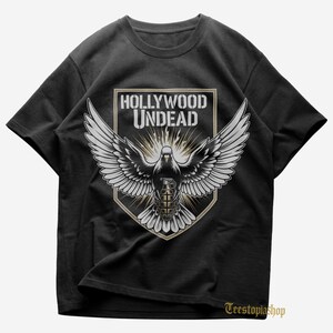 May include: Black t-shirt featuring a graphic of a dove with outstretched wings, a grenade body, and the text "HOLLYWOOD UNDEAD" in a shield design. The shirt has a crew neck and short sleeves. The brand name "Teestopiashop" is printed at the bottom right.