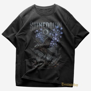 May include: Black t-shirt featuring a detailed graphic of a raven with mechanical elements and butterfly wings, set against a dark, atmospheric background. The band name "SHINEDOWN" is displayed across the top. The shirt has a crew neck and short sleeves.