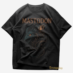 May include: Black t-shirt featuring the band name "MASTODON" in brown letters above a detailed illustration. The illustration depicts a teal dragon-like creature with orange eyes and tentacles, with a steampunk-style structure on its back.