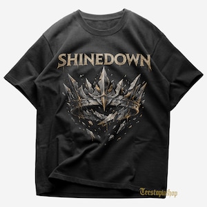 May include: Black t-shirt featuring the band name "SHINEDOWN" in gold lettering above a detailed crown design. The crown is silver and gold with a shattered, textured appearance. The shirt has a crew neck and short sleeves.