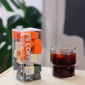 May include: A clear plastic box filled with orange, beige, and black coffee cubes, with the text "F5 COFFEE MIX" printed on the side. A glass of iced coffee sits next to the box on a light-colored tray.