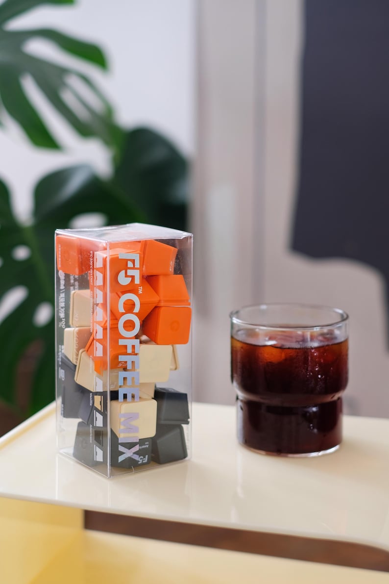 May include: A clear plastic box filled with orange, beige, and black coffee cubes, with the text "F5 COFFEE MIX" printed on the side. A glass of iced coffee sits next to the box on a light-colored tray.