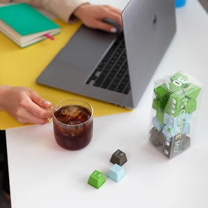 Pu&ograve; includere: Una scena di lavoro con un laptop, un bicchiere di caff&egrave; freddo e una scatola trasparente di tasti della tastiera verdi, blu e marroni. Sono visibili anche un quaderno verde e carta gialla. I tasti sono etichettati "F5 COFFEE MIX".
