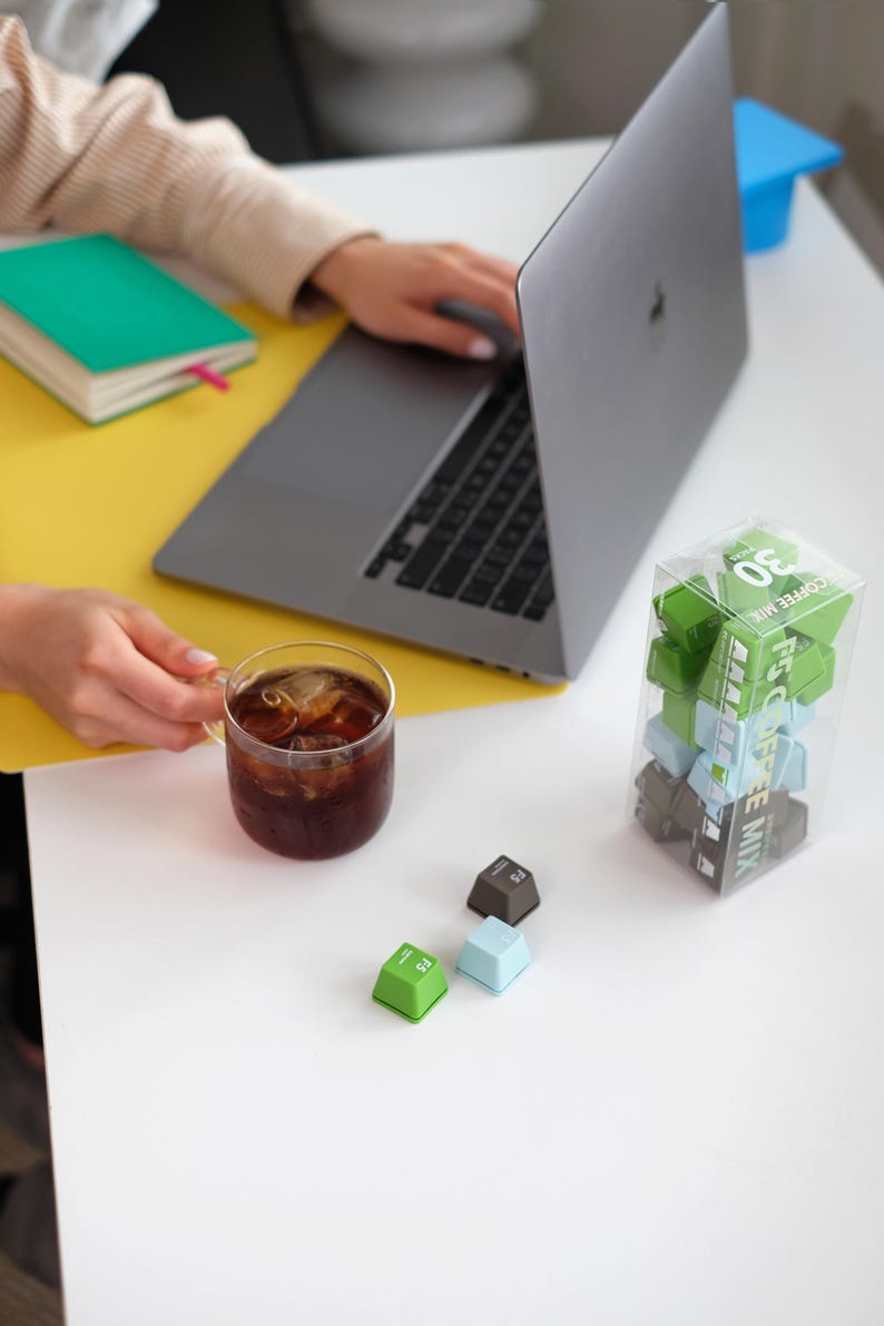 Pu&ograve; includere: Una scena di lavoro con un laptop, un bicchiere di caff&egrave; freddo e una scatola trasparente di tasti della tastiera verdi, blu e marroni. Sono visibili anche un quaderno verde e carta gialla. I tasti sono etichettati "F5 COFFEE MIX".