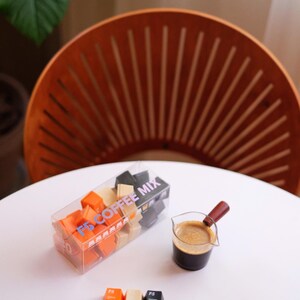 May include: A clear box of F5 Coffee Mix cubes, with orange, beige, and black cubes. A small glass with a wooden handle holds coffee. Three F5 keycaps are on the table. The scene is on a white table.