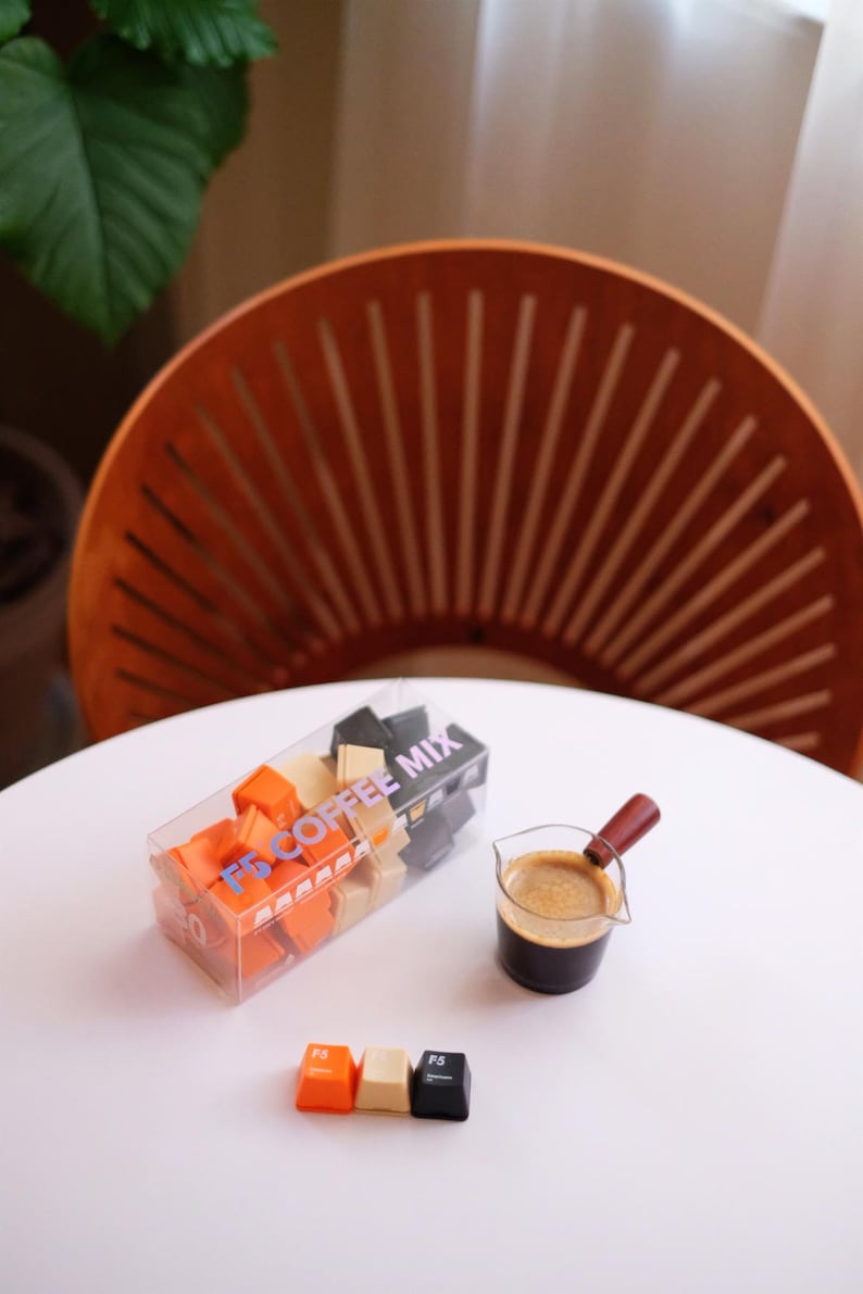 May include: A clear box of F5 Coffee Mix cubes, with orange, beige, and black cubes. A small glass with a wooden handle holds coffee. Three F5 keycaps are on the table. The scene is on a white table.
