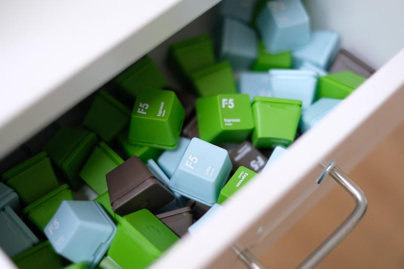 Pu&ograve; includere: Un cassetto pieno di tasti F5 colorati in tonalit&agrave; di verde, blu e marrone. Ogni tasto ha il testo "F5" e "Espresso" stampato sopra. Il cassetto &egrave; bianco con una maniglia argentata. I tasti sono in diverse tonalit&agrave;.