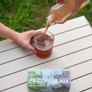 Pu&ograve; includere: Un bicchiere di t&egrave; freddo viene versato da una bottiglia. Il bicchiere &egrave; su un tavolo bianco accanto a una scatola trasparente di tasti per tastiera F5 COFFEE MIX in verde, blu e grigio.