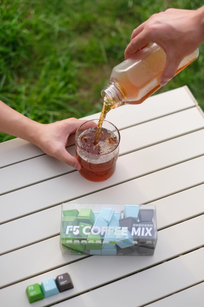 Pu&ograve; includere: Un bicchiere di t&egrave; freddo viene versato da una bottiglia. Il bicchiere &egrave; su un tavolo bianco accanto a una scatola trasparente di tasti per tastiera F5 COFFEE MIX in verde, blu e grigio.