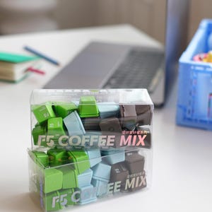 Pu&ograve; includere: Due contenitori di plastica trasparente pieni di tasti quadrati colorati. I tasti sono verdi, azzurri e marroni. I contenitori hanno la scritta "F5 COFFEE MIX". Un laptop e altri articoli per l'ufficio sono sullo sfondo.