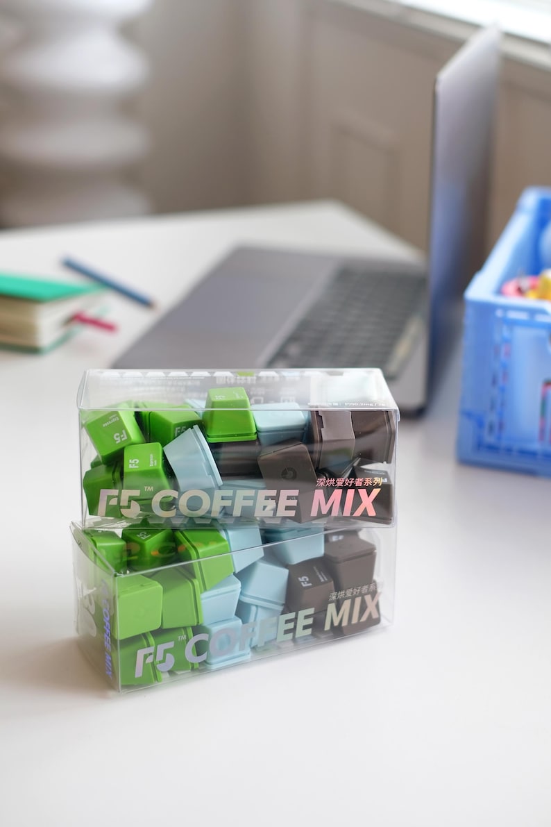 Pu&ograve; includere: Due contenitori di plastica trasparente pieni di tasti quadrati colorati. I tasti sono verdi, azzurri e marroni. I contenitori hanno la scritta "F5 COFFEE MIX". Un laptop e altri articoli per l'ufficio sono sullo sfondo.