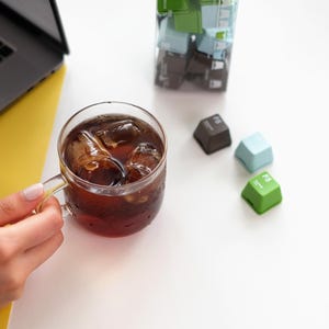 Pu&ograve; includere: Una tazza di vetro piena di caff&egrave; freddo, tenuta in mano. Tre tasti colorati della tastiera sono sparsi: marrone, azzurro e verde. Un computer portatile e una superficie gialla sono sullo sfondo.