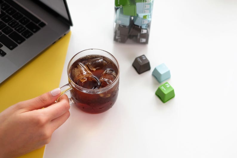 Pu&ograve; includere: Una tazza di vetro piena di caff&egrave; freddo, tenuta in mano. Tre tasti colorati della tastiera sono sparsi: marrone, azzurro e verde. Un computer portatile e una superficie gialla sono sullo sfondo.