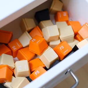 May include: A collection of colorful F5 keycaps in a white drawer. The keycaps are in shades of orange, cream, and black. Each keycap has the text "F5" printed on it. The drawer is slightly open, revealing the contents.