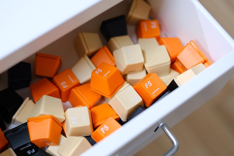 May include: A collection of colorful F5 keycaps in a white drawer. The keycaps are in shades of orange, cream, and black. Each keycap has the text "F5" printed on it. The drawer is slightly open, revealing the contents.