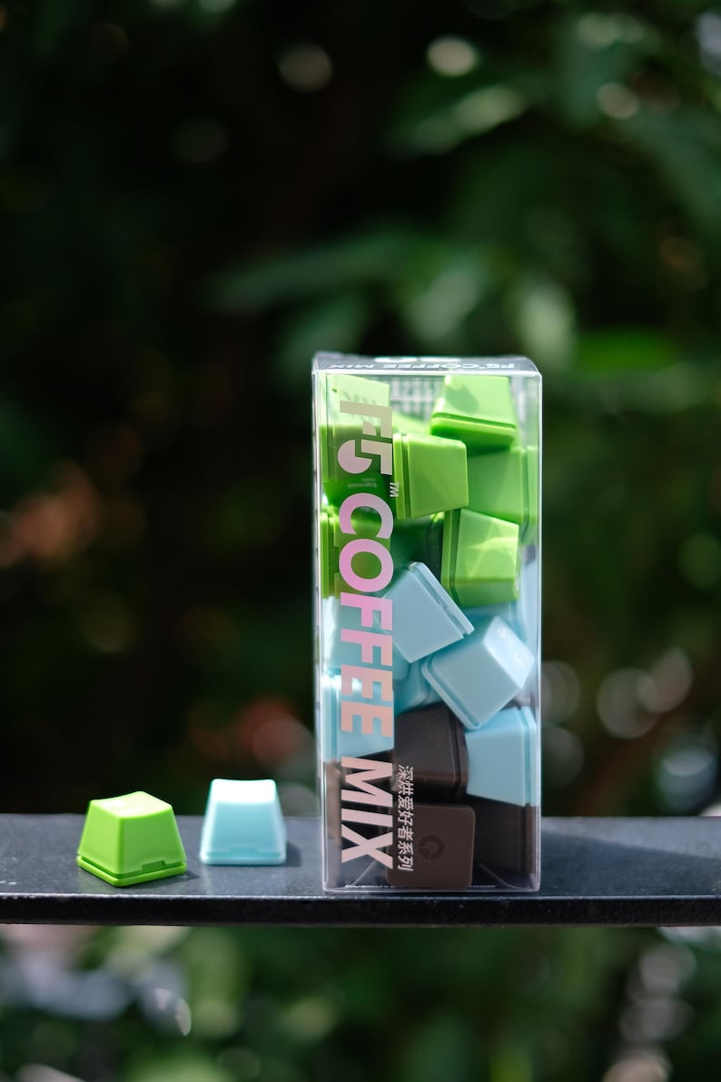 Pu&ograve; includere: Una scatola trasparente piena di cubetti di miscela di caff&egrave; quadrati e colorati, nei toni del verde, blu e marrone. La scatola ha la scritta "T5 COFFEE MIX". Due cubetti sono fuori dalla scatola.