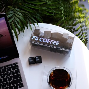 May include: A clear plastic box filled with black square objects, labeled "F5 COFFEE", sits on a white surface next to a laptop and a glass of coffee. Two "F5" keycaps are also present. Green foliage is in the background.