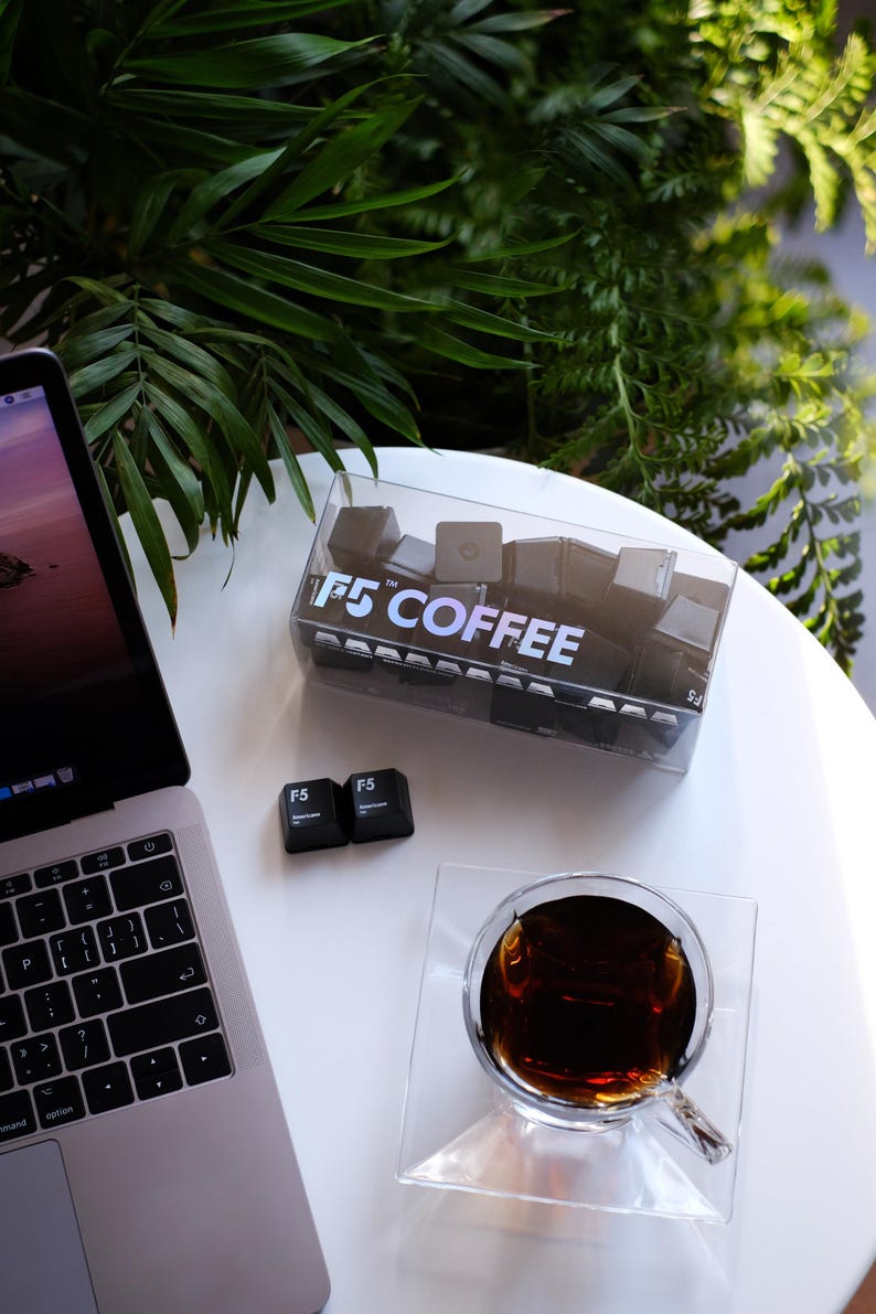 May include: A clear plastic box filled with black square objects, labeled "F5 COFFEE", sits on a white surface next to a laptop and a glass of coffee. Two "F5" keycaps are also present. Green foliage is in the background.