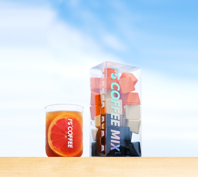 May include: A glass of iced tea with a grapefruit slice and a clear box of coffee mix cubes. The box has the text "F5 COFFEE MIX" in a vertical design. The glass also has the text "F5 COFFEE". The background is a bright blue sky.