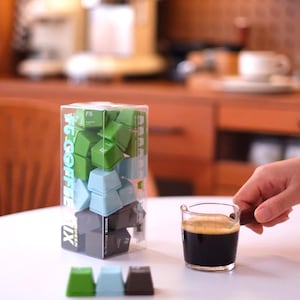 Pu&ograve; includere: Un contenitore di plastica trasparente pieno di tasti verdi, blu e grigi, etichettato "#6 COFFEE MIX". Un piccolo bicchiere di espresso &egrave; tenuto in mano. Tre tasti sono disposti su una superficie bianca.