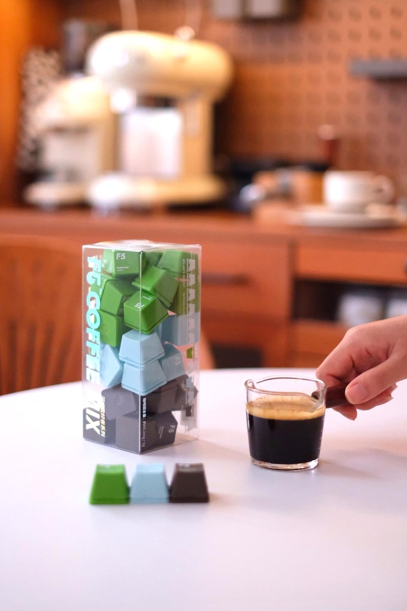Pu&ograve; includere: Un contenitore di plastica trasparente pieno di tasti verdi, blu e grigi, etichettato "#6 COFFEE MIX". Un piccolo bicchiere di espresso &egrave; tenuto in mano. Tre tasti sono disposti su una superficie bianca.