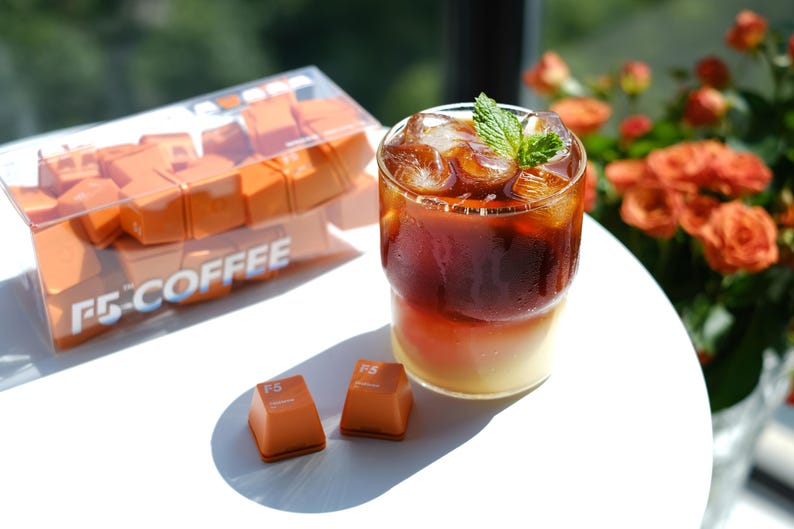 May include: A refreshing iced coffee drink with a sprig of mint, served in a clear glass. Beside it are two orange-colored, square-shaped candies and a box of similar candies. The box has the text "F5 COFFEE".