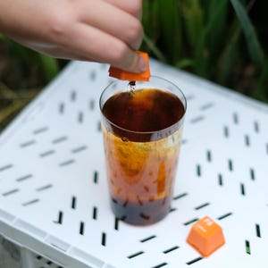 May include: A tall glass of dark liquid with an orange square being dropped into it. The drink is on a white surface with a second orange square nearby. The background is blurred green foliage.