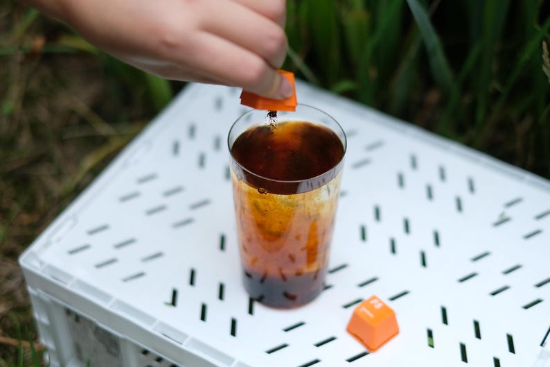 May include: A tall glass of dark liquid with an orange square being dropped into it. The drink is on a white surface with a second orange square nearby. The background is blurred green foliage.