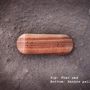 Könnte beinhalten: Ein Fingerboard aus rötlich-braunem Holz mit einem Santos-Pallisander-Boden. Das Fingerboard hat eine abgerundete Form und kleine Löcher zur Montage. Der Text "Top; Vtec red" und "Bottom; Santos pallisander" ist sichtbar.