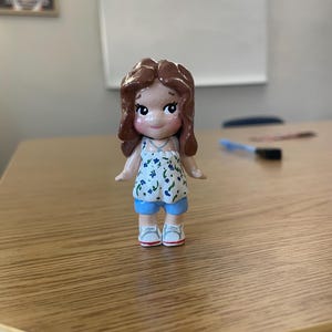 May include: A handcrafted figurine of a girl with brown hair, wearing a white top with blue floral patterns, blue shorts, and white sneakers. The doll has rosy cheeks and a sweet expression. The figurine is standing on a wooden surface.