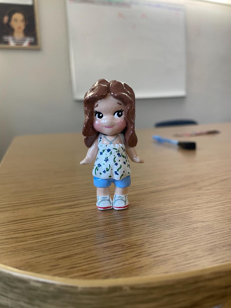 May include: A handcrafted figurine of a girl with brown hair, wearing a white top with blue floral patterns, blue shorts, and white sneakers. The doll has rosy cheeks and a sweet expression. The figurine is standing on a wooden surface.