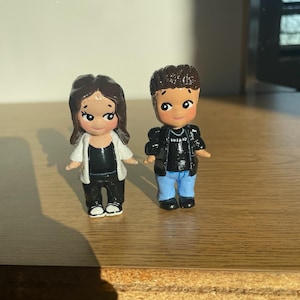 May include: Two small, hand-painted figurines of a couple. The female figure wears a black top, black pants, and a white jacket. The male figure wears a black jacket, blue jeans, and a black shirt. Both have dark hair and are standing on a wooden surface.