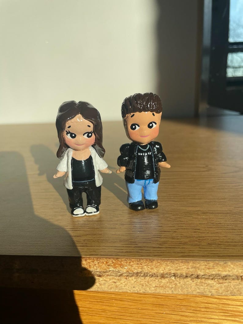 May include: Two small, hand-painted figurines of a couple. The female figure wears a black top, black pants, and a white jacket. The male figure wears a black jacket, blue jeans, and a black shirt. Both have dark hair and are standing on a wooden surface.