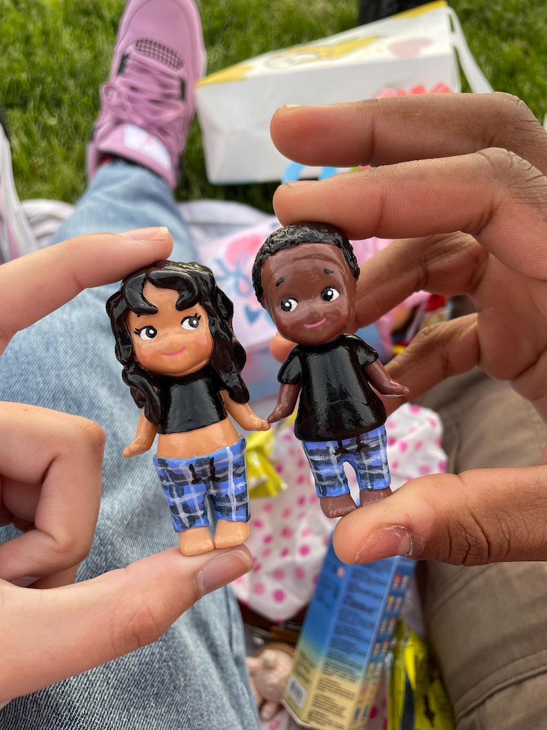 May include: Two small, hand-painted dolls are held up. One doll has dark hair and the other has short black hair. Both dolls wear black tops and blue plaid pants. The dolls have rosy cheeks and are smiling.