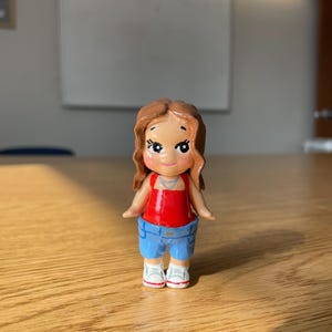 May include: A small, hand-painted figurine of a person. The figure has brown hair, a red top, blue shorts, and white sneakers. The figurine is standing on a wooden surface.