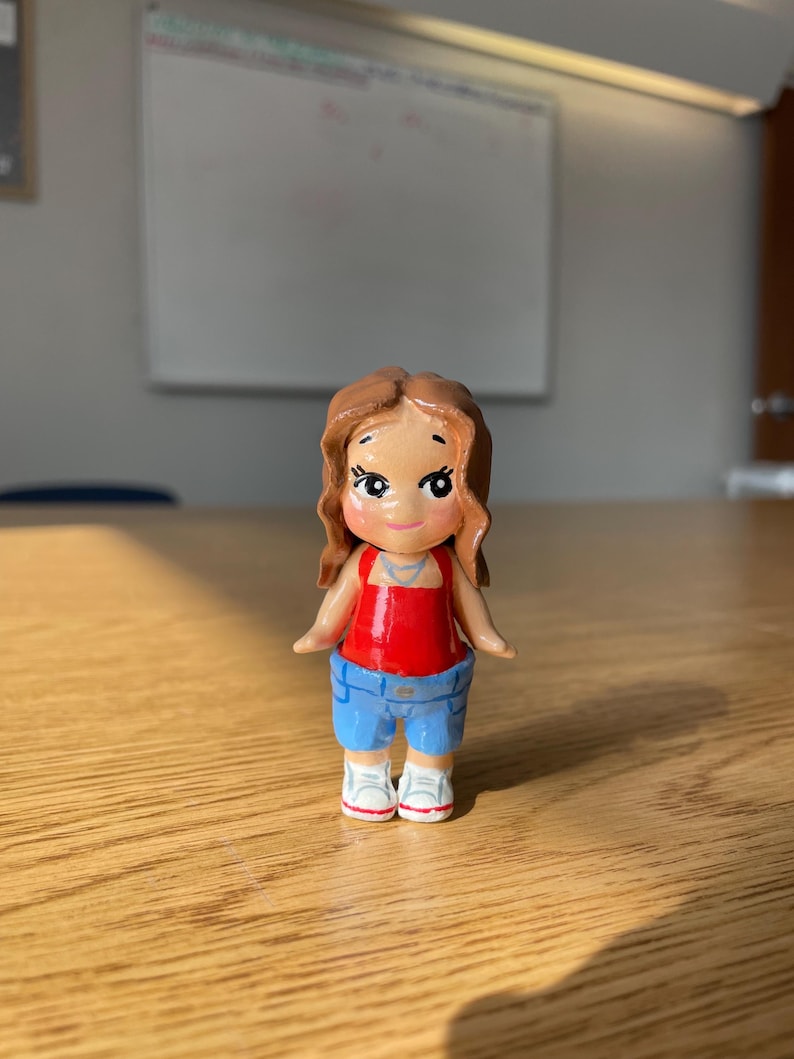 May include: A small, hand-painted figurine of a person. The figure has brown hair, a red top, blue shorts, and white sneakers. The figurine is standing on a wooden surface.