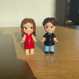 May include: Two small, hand-painted figurines of a couple. The woman wears a red dress and has brown hair. The man is in a black shirt and blue pants. Both have smiling faces and are standing on a wooden surface. The figurines are likely handmade.