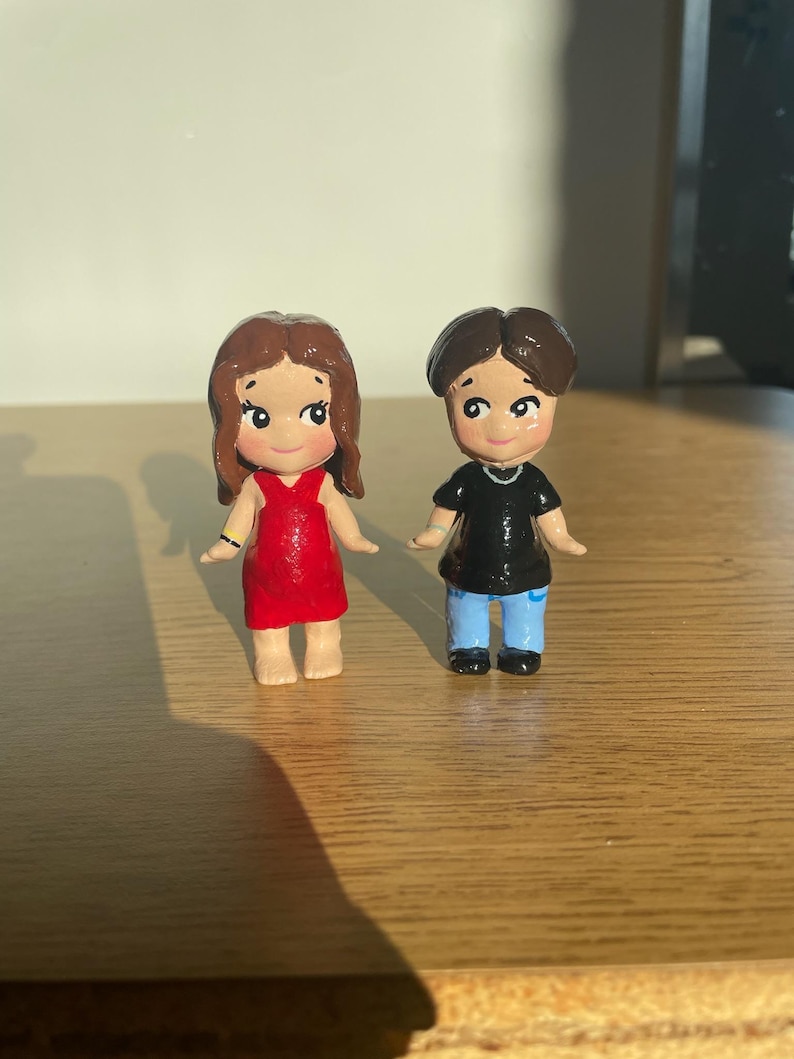 May include: Two small, hand-painted figurines of a couple. The woman wears a red dress and has brown hair. The man is in a black shirt and blue pants. Both have smiling faces and are standing on a wooden surface. The figurines are likely handmade.